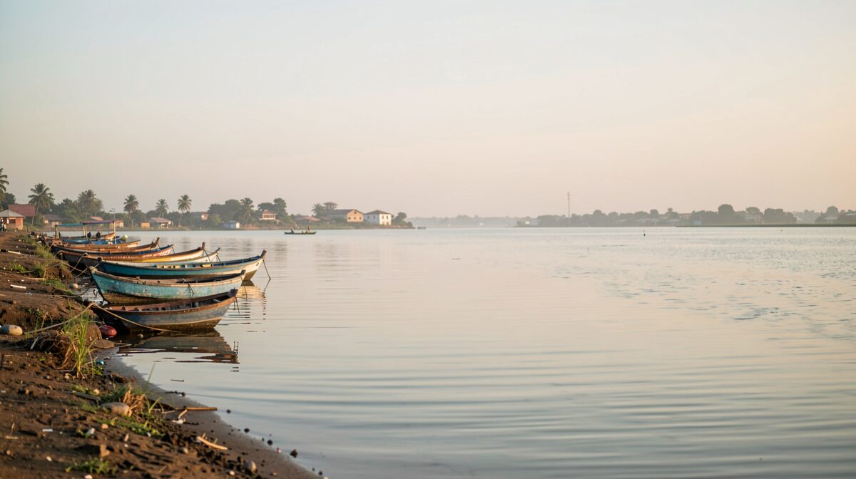 A calm stretch of the Ébrié lagoon in Abidjan at early morning, boats moored along the shore, soft golden light, wide horizontal framing.